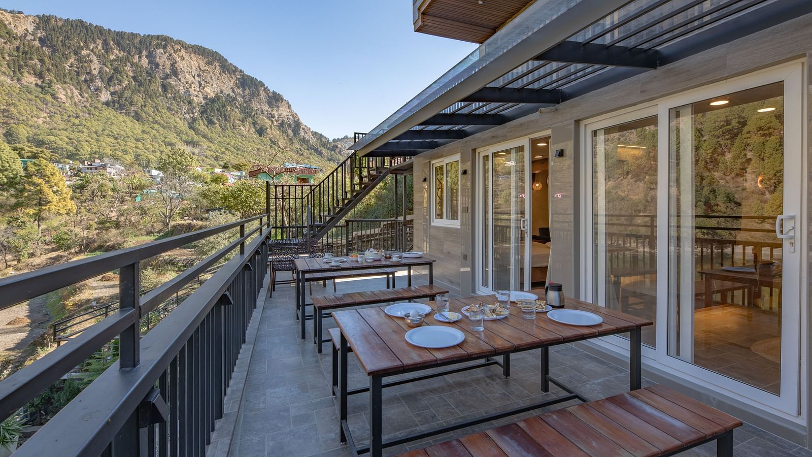 Outdoor dining tables set on a narrow balcony with views of green hills and a clear sky.