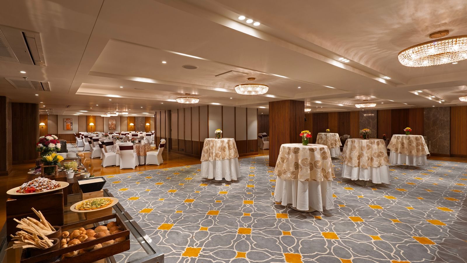 This image depicts a spacious and elegant banquet hall, The room features high ceilings, ornate chandeliers, and a plush carpeted floor with several round tables are arranged throughout the space.