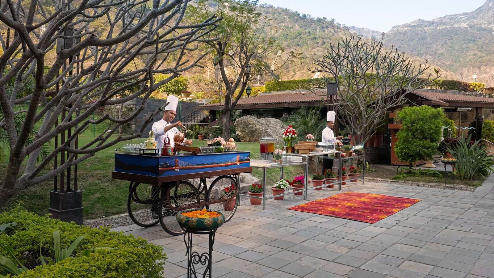 Two chefs are preparing food at outdoor food stations. There are mountains in the background and lush surrounding the scene.