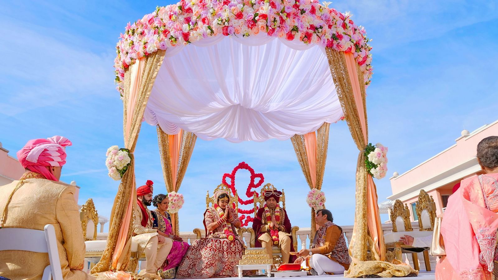 This image showcases a grand Indian wedding ceremony taking place outdoors under a beautiful, floral-adorned setup. 