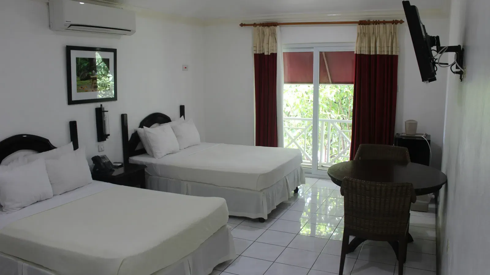 A spacious hotel room contains two beds with white bedding, a dark dresser with a television, and red curtains | Deluxe Double Room | Altamont Court, Jamaica