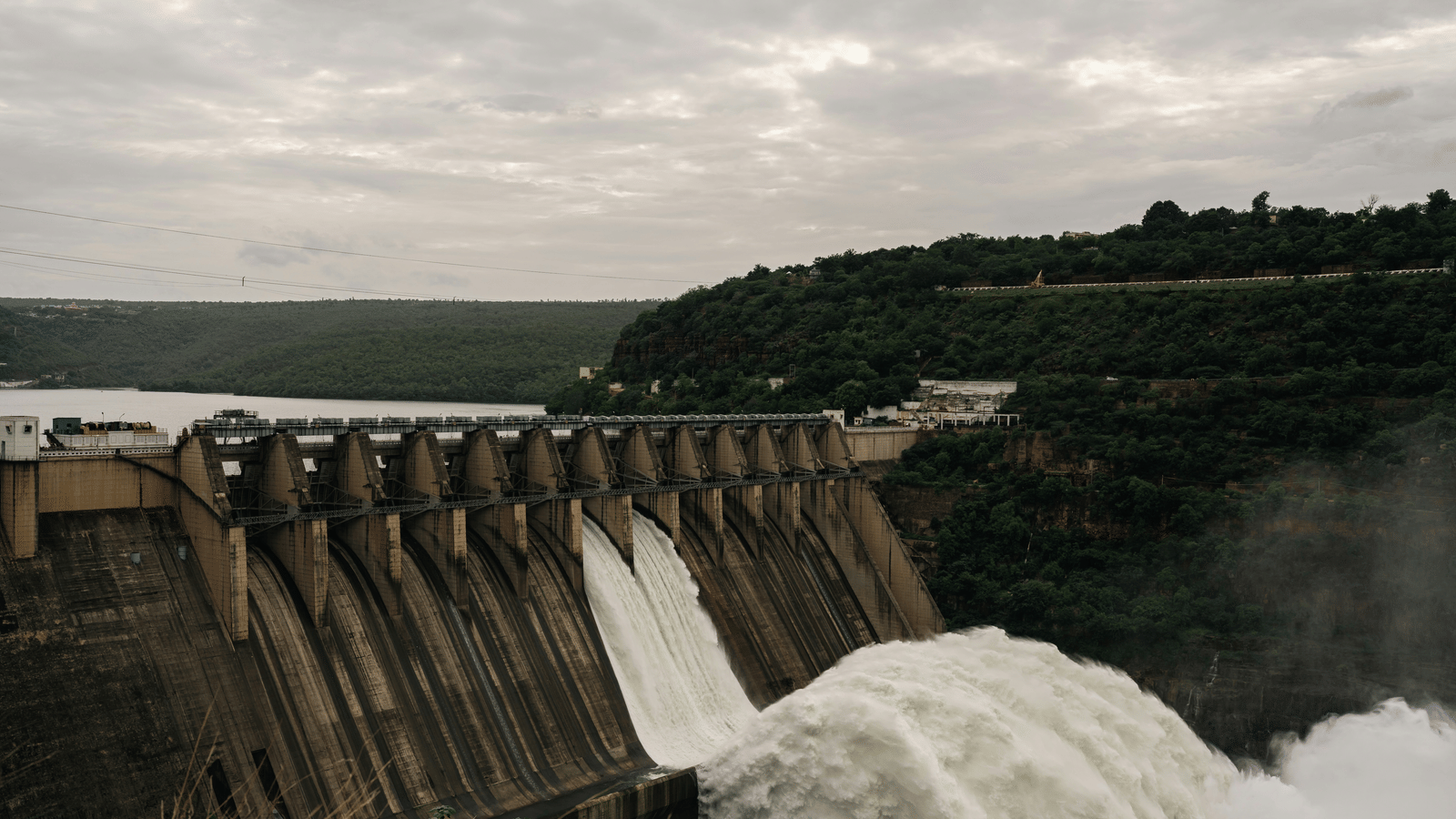 A massive dam with multiple sluice gates open, powerfully releasing a huge volume of white water into a river below.