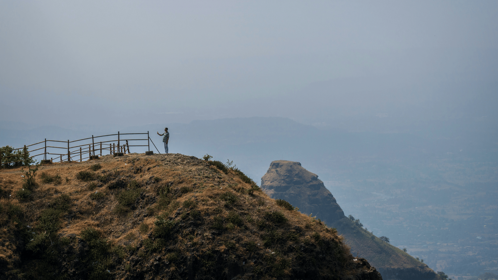 A hazy, wide landscape shot from a dry hilltop viewpoint with a small wooden fence, overlooking layered mountains fading into the distance.