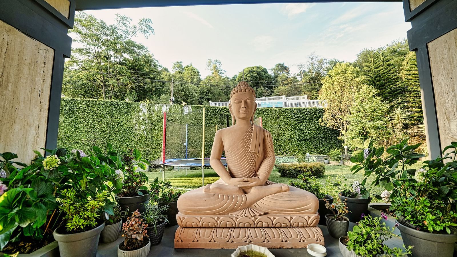 A picture of a statue placed at the centre, captured in the day light and surrounded with few plants.
