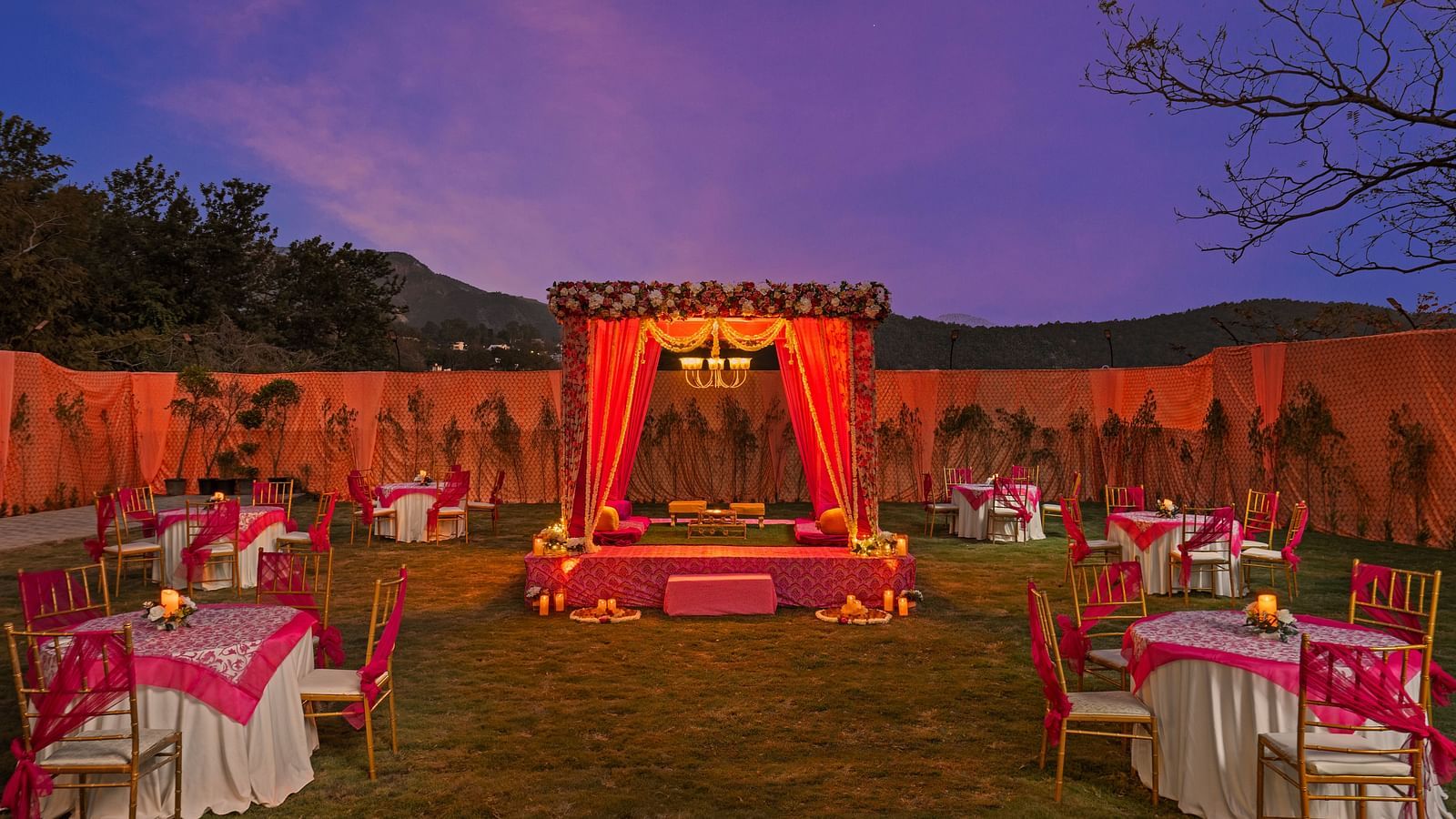  Outdoor evening wedding setup with lit mandap, pink tablecloths, and chairs, creating a romantic, twilight ambiance.