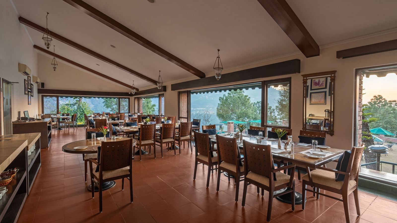 A restaurant with glass windows offering a beautiful mountain view, featuring dining tables arranged with chairs, a mirror on the wall, and stylish pendant lights overhead -  Baikunth resort, kasauli 