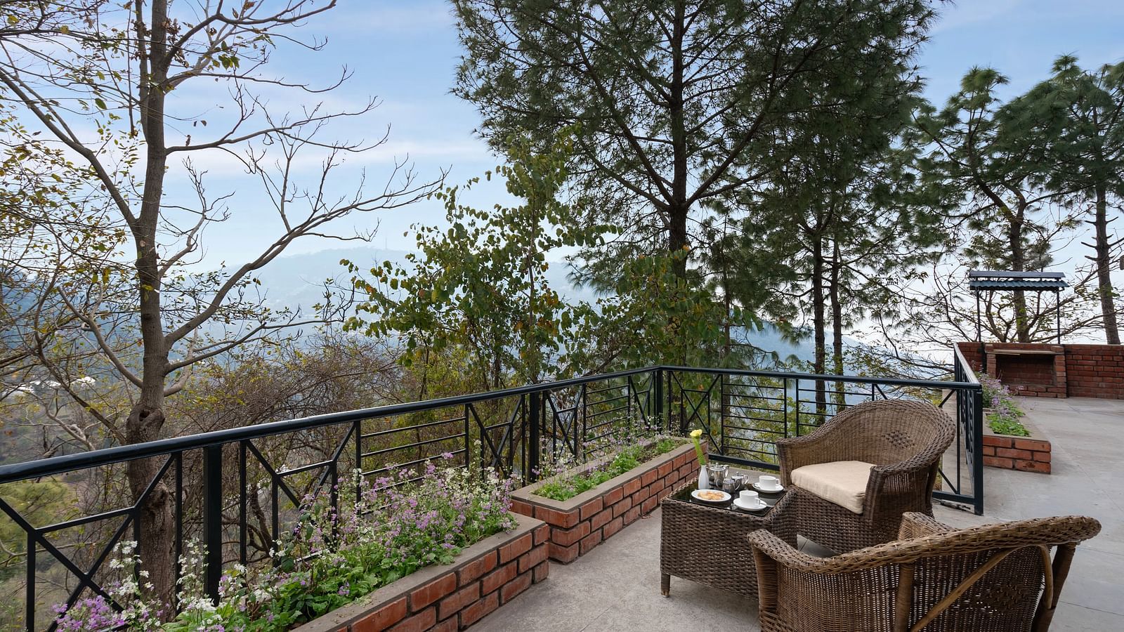 Two wooden chairs and a table set on an open balcony with a view of trees and mountains afar - Baikunth resort, kasauli 