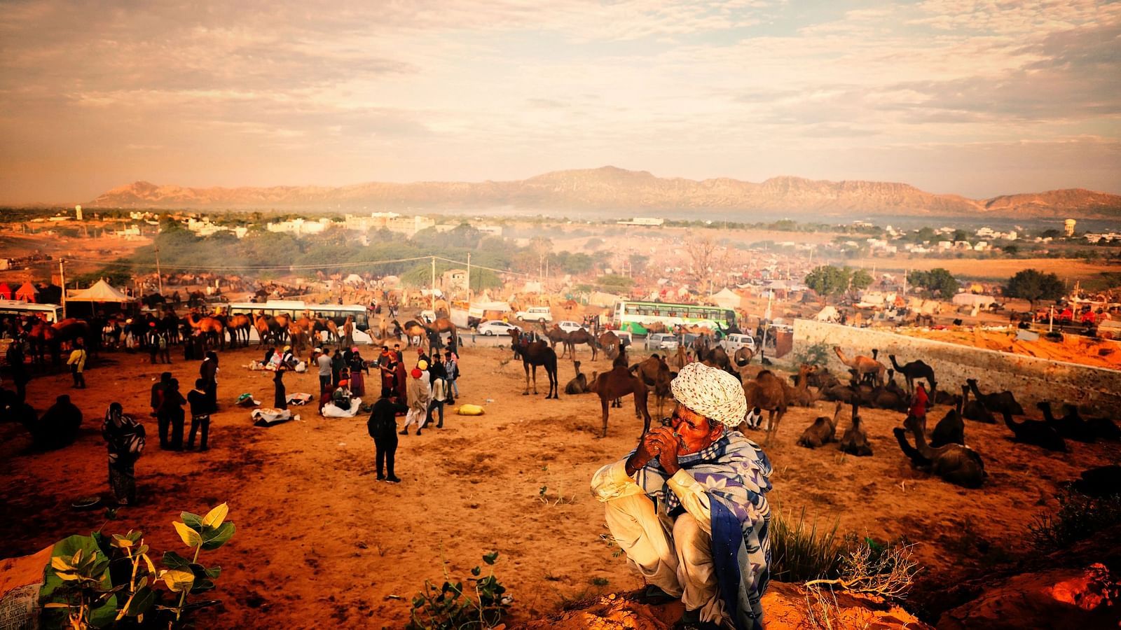 A wide, dusty landscape with distant hills, filled with people and animals.