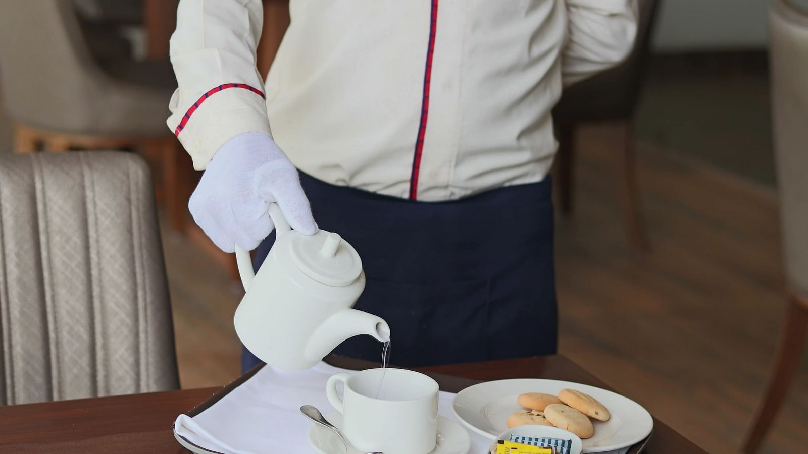 A waiter in a uniform pours a drink from a teapot into a cup on a tray. - Bamboo Saa, Udaipur
