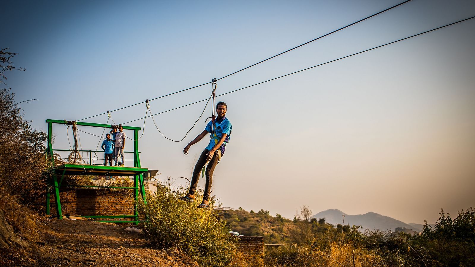 A person zip-lining across a scenic landscape with hills in the background.- Bamboo Saa Resort & Spa, Udaipur