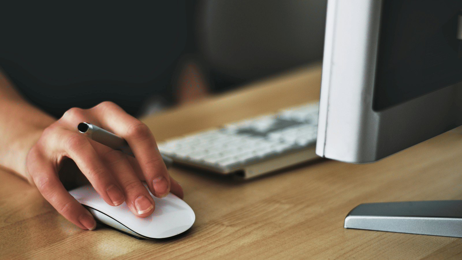 A person's hand on a computer mouse, working at a desk with a keyboard and monitor.