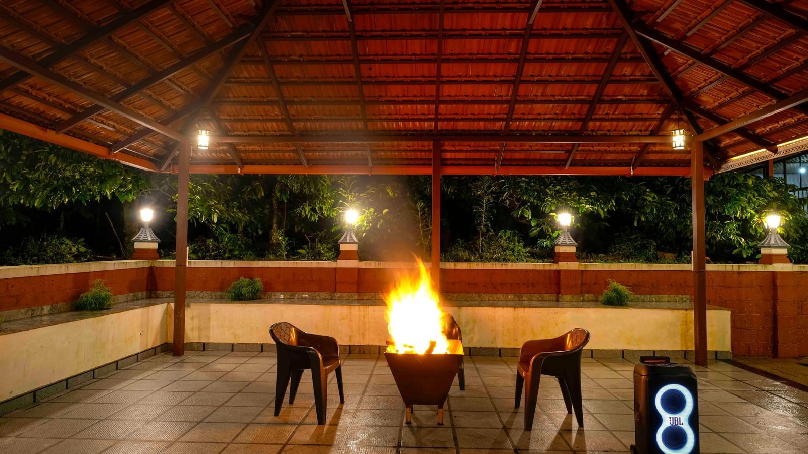 A large bonfire crackles at the center of a covered, open-air pavilion at Bhuthanakadu Retreat, surrounded by chairs under the warm glow of the lights at night.