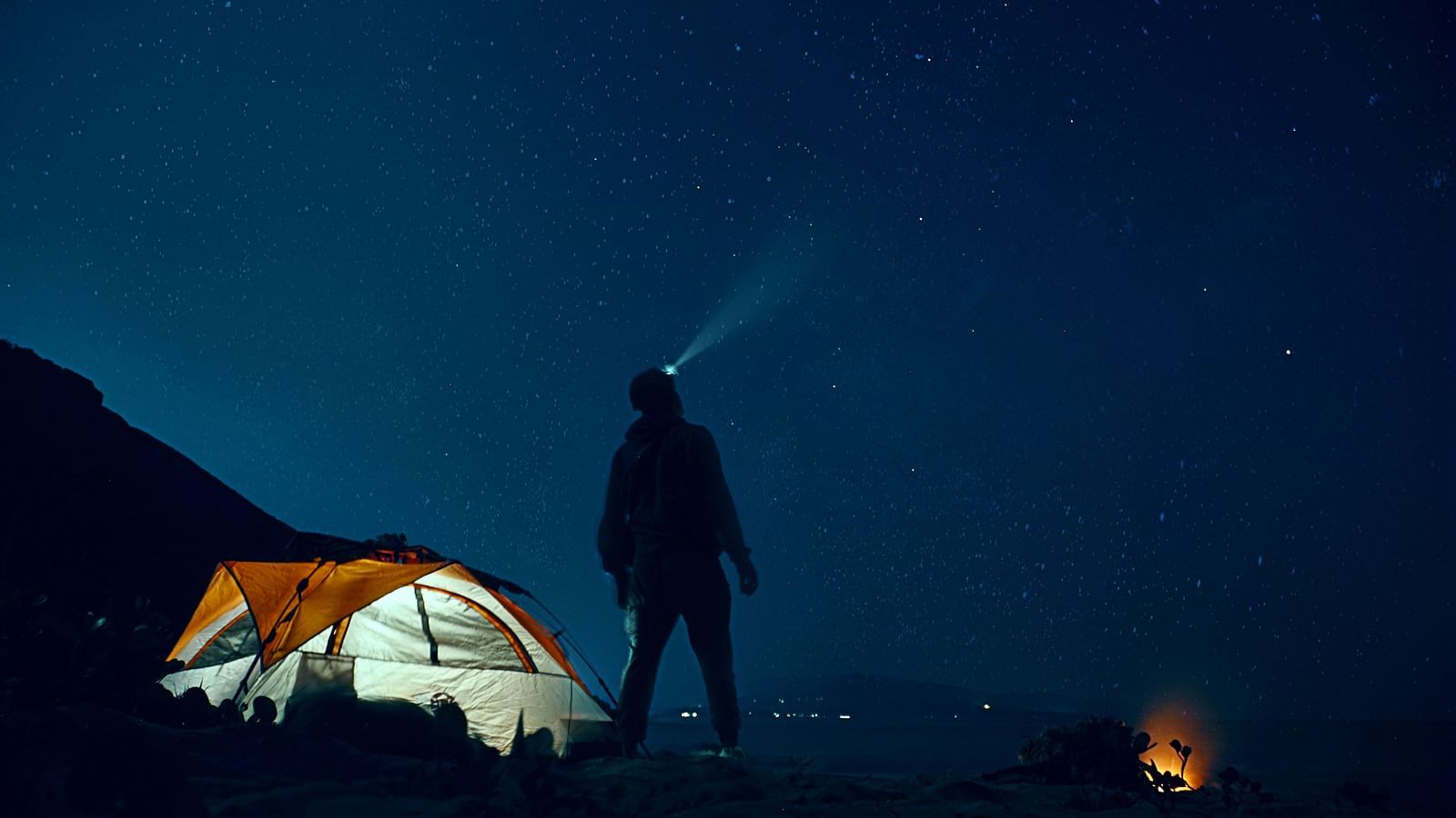 A person standing near a glowing tent, gazing at the night sky