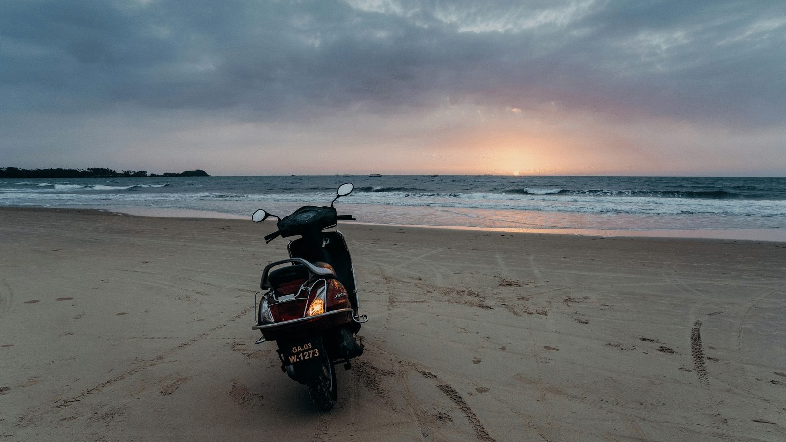A scooter is parked on a beach at sunset with a dramatic cloudy sky.
