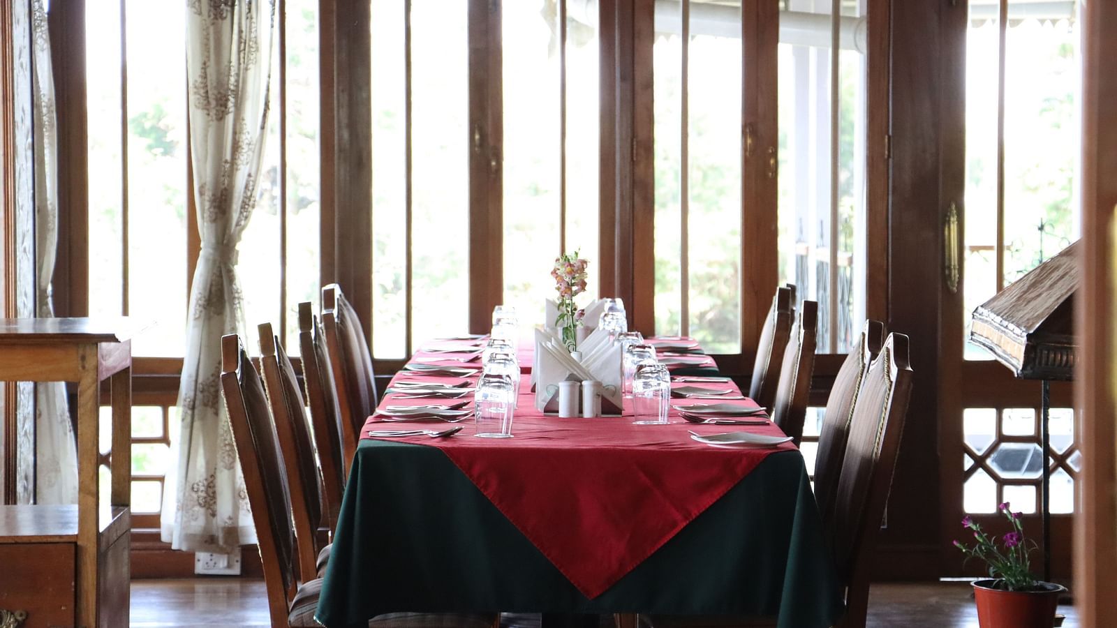 The eight seating table in the dining room of Cedar Inn, Darjeeling placed beside a glass and wood framed wall, arranged with cutlery and other dining amenities.
