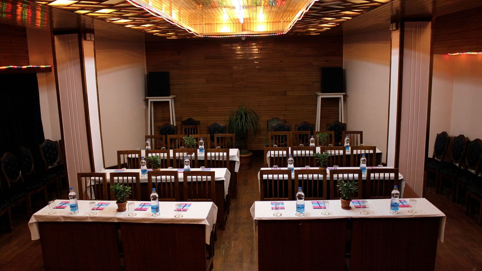 Classroom seating arrangement in the Banquet hall of Cedar Inn, Darjeeling featuring six tables with four chairs each, overhead lighting and speakers.