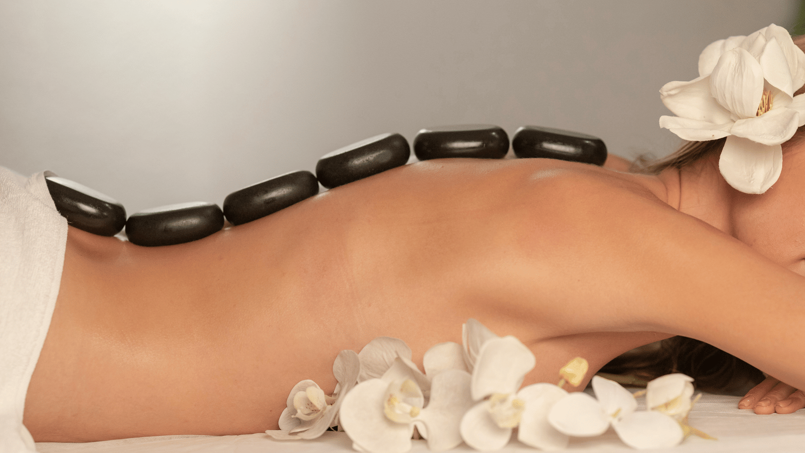 Polished black massage stones are lined up along a woman's back as she lies on a spa bed decorated with white flowers.