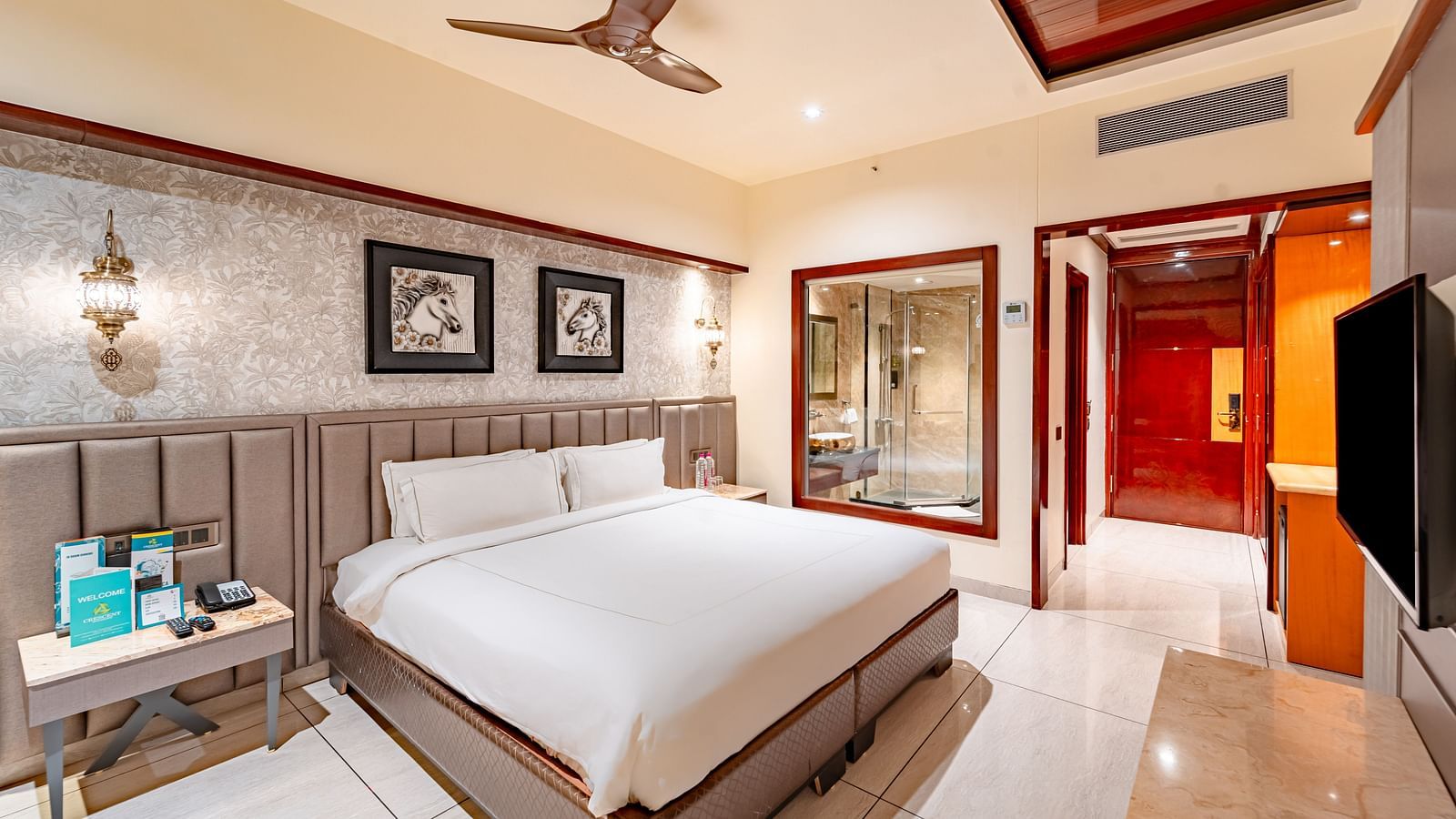 A spacious bed with two photo frames above, a bedside table with essentials, and a wall mounted TV opposite at  Crescent Spa & Resort, Indore