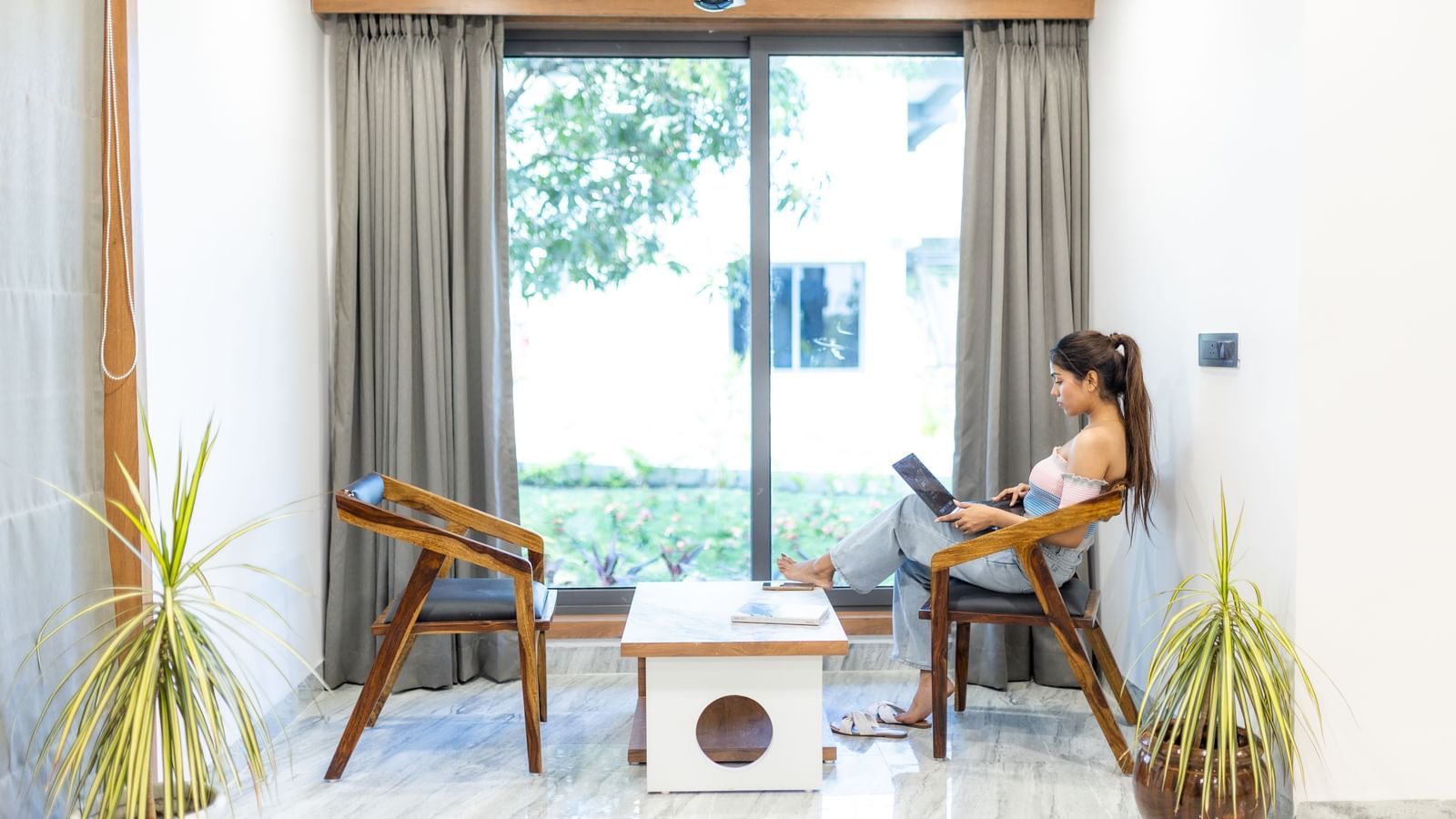 A woman sitting in the seating area for 2, located by the large glass windows with curtains.