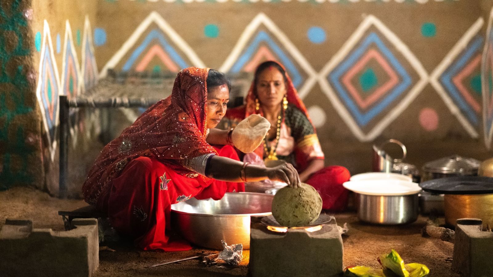 Two woman seated on the floor, preparing food using bit steel utensils in front of a cot placed in a room that has traditional wall markings -Daksh Eden Greenz, Sasan Gir