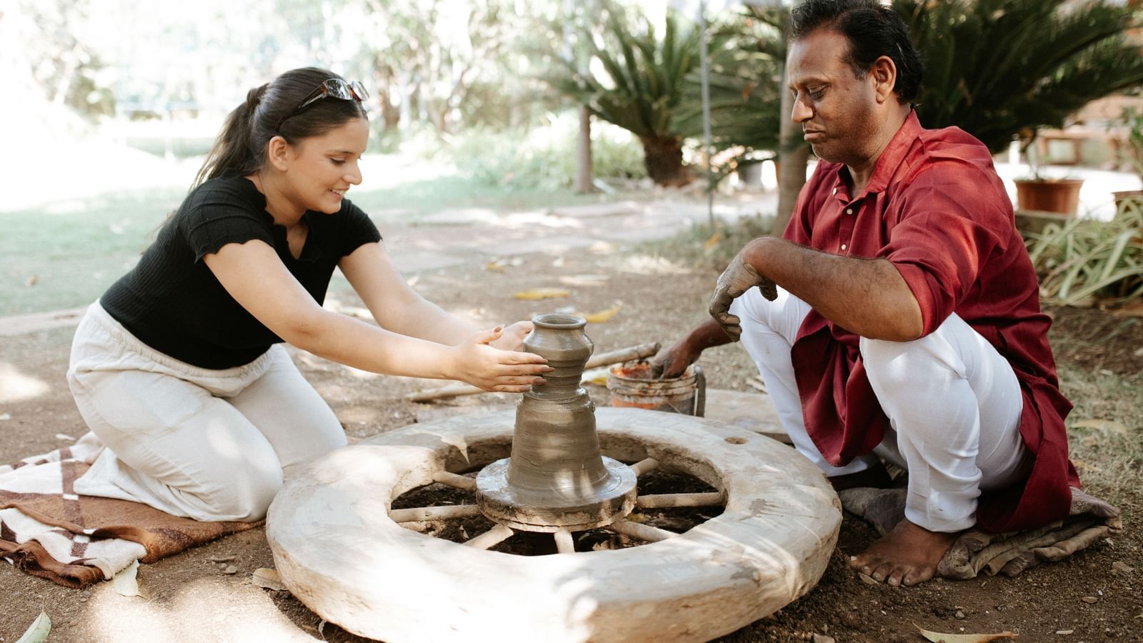 A woman moulding clay on a pottery wheel while a man watches, seated on the ground at Daksh Eden Greenz, Sasan Gir.
