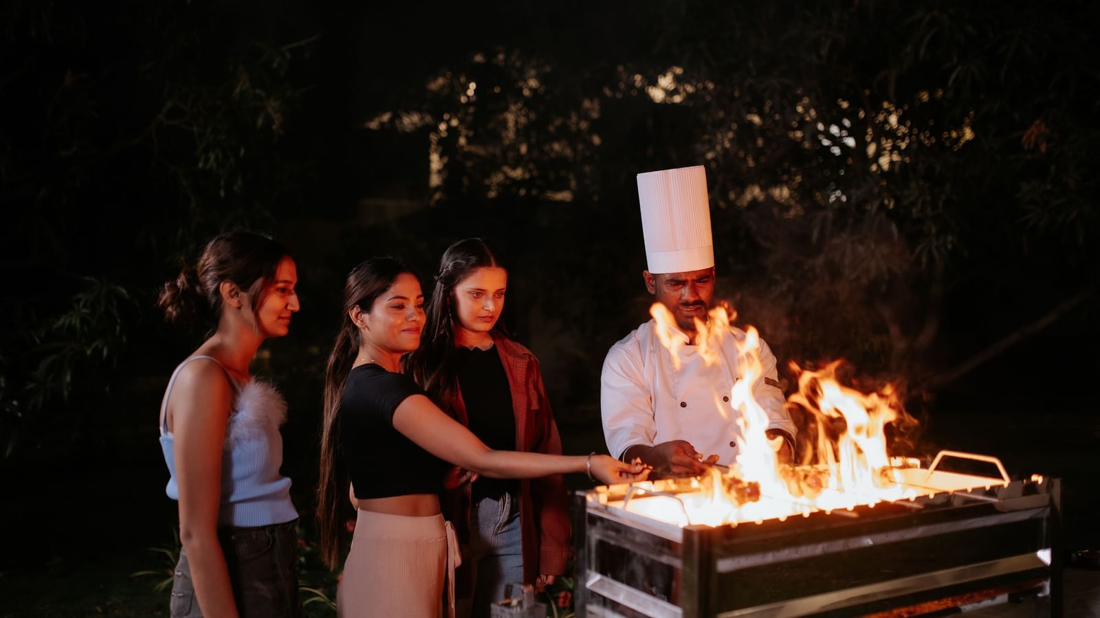 a chef cooking food at the resort with three other people on a fire grill at night -Daksh Eden Greenz, Sasan Gir
