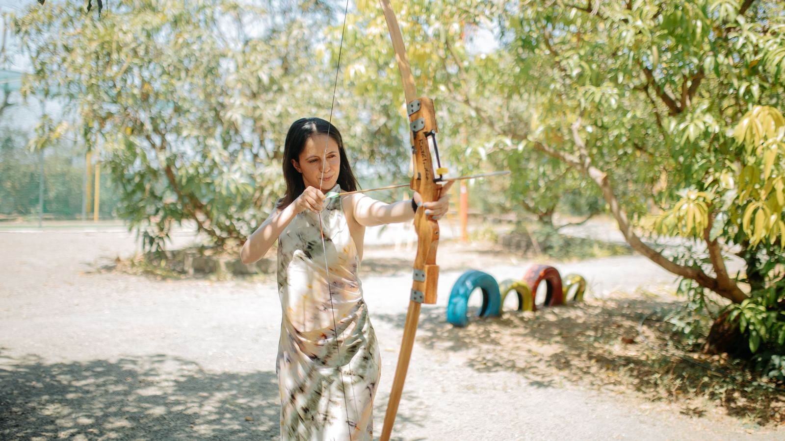 A woman holding a bow in an archery stance, appearing ready to shoot, amidst trees at Daksh Resort & Amusement Park.