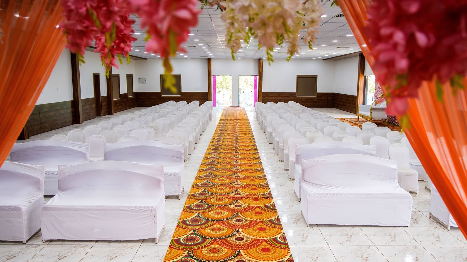 A close up of a hall at Daksh Resort & Amusement Park featuring arranged chairs and a patterned isle with decorated curtains on the foreground.
