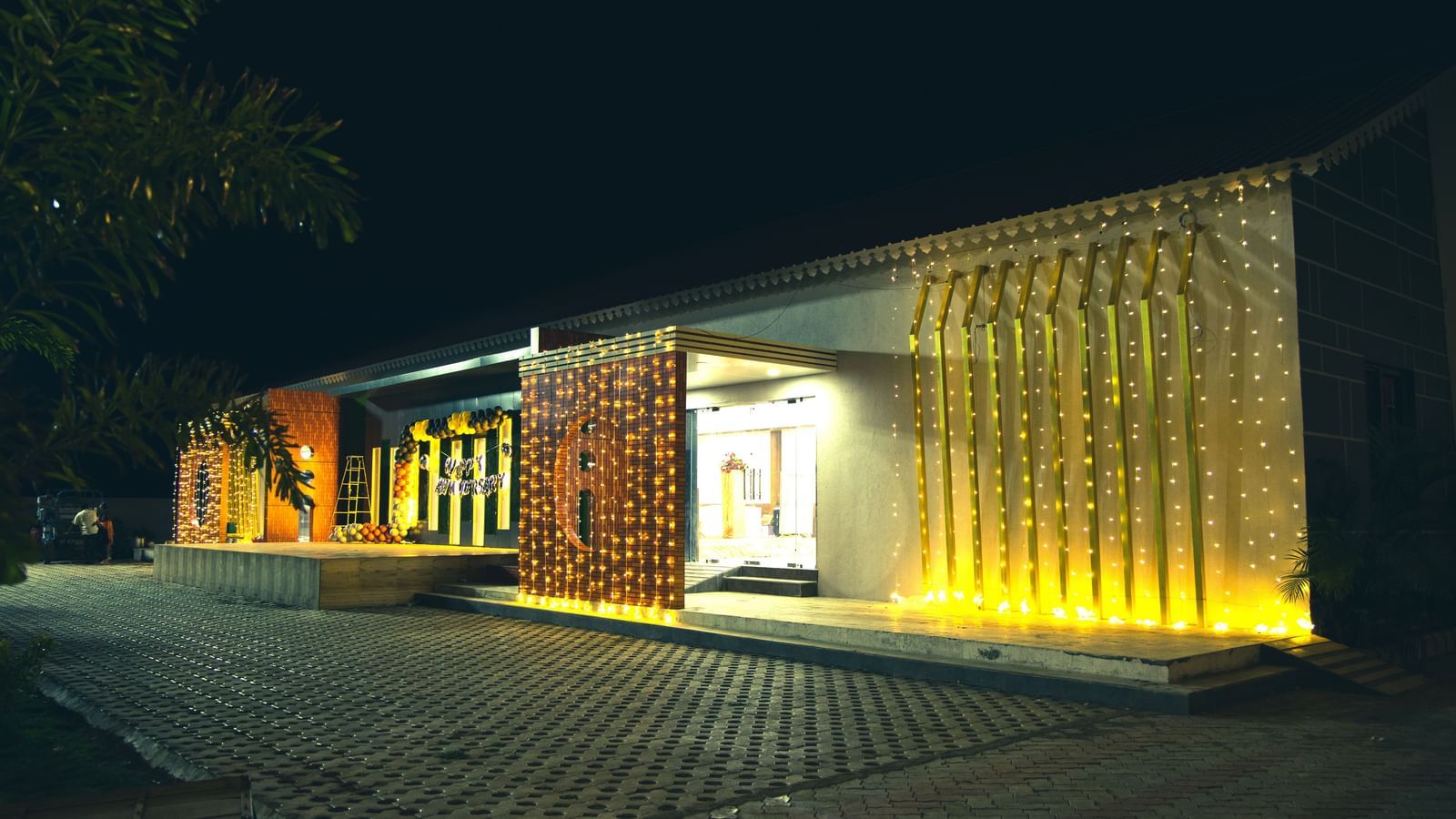 A night view of the entry of an event space in Daksh Eden Greenz decorated with fairy lights and a 'happy birthday' balloon arrangement on the wall.