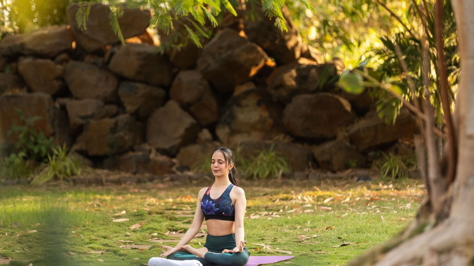 A woman practising yoga amidst the greenery and a low height rock wall at the background at Daksh Resort & Amusement Park.