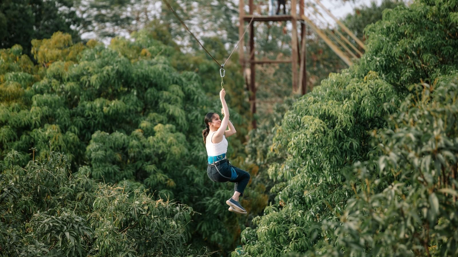 A person zip-lining through a dense green forest area, suspended mid-air by harness and cable at Daksh Resort & Amusement Park.