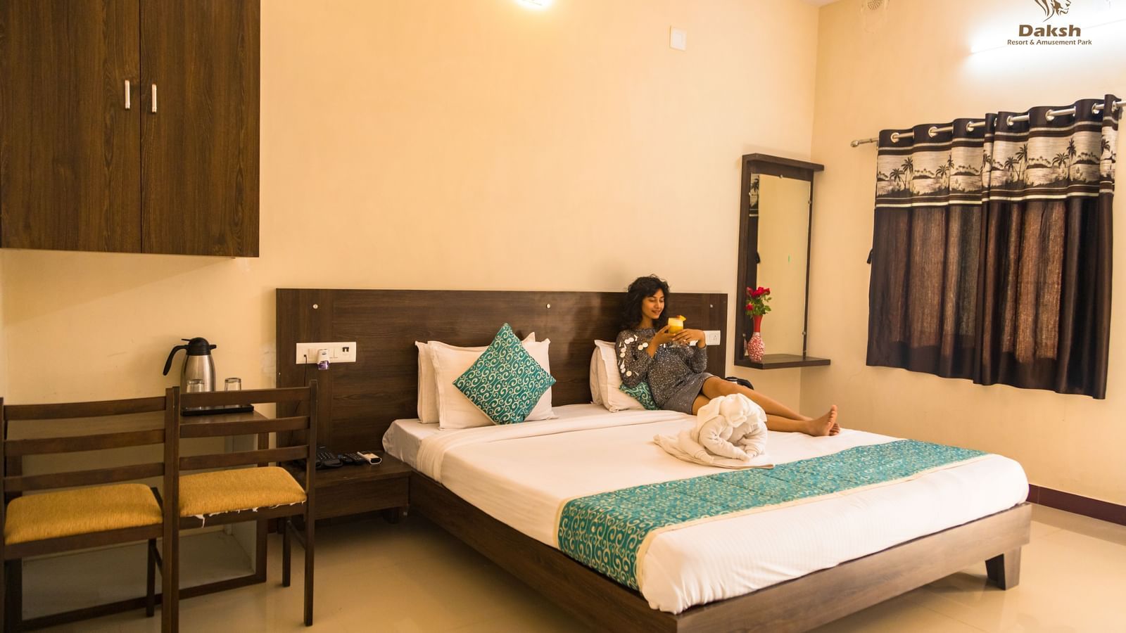 A guest room with a double bed, chairs, and dressing area, with a woman seated at Daksh Resort & Amusement Park.