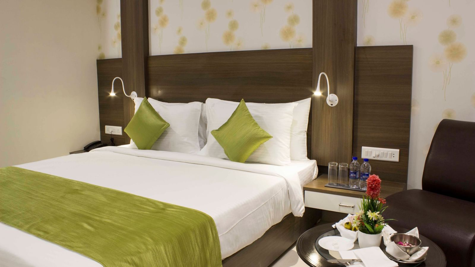 A hotel bedroom with a neatly made bed, green accents, and bedside tables with lamps | Daksh Comfort Room | Daksh The Madhuvan Suites