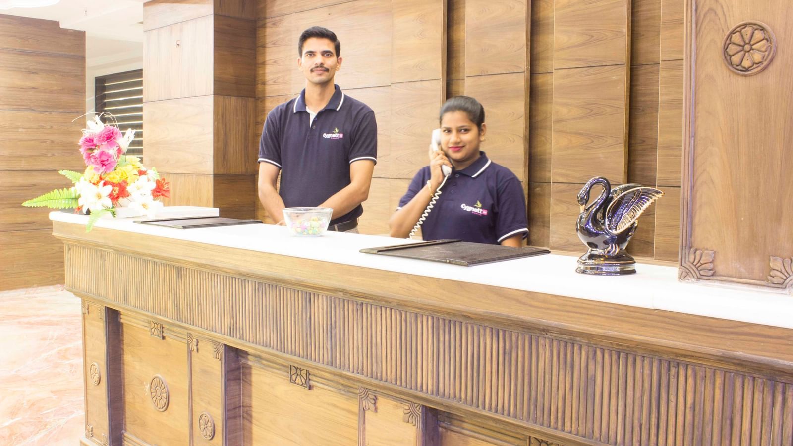 A hotel reception desk with two staff members smiling | Daksh The Madhuvan Suites