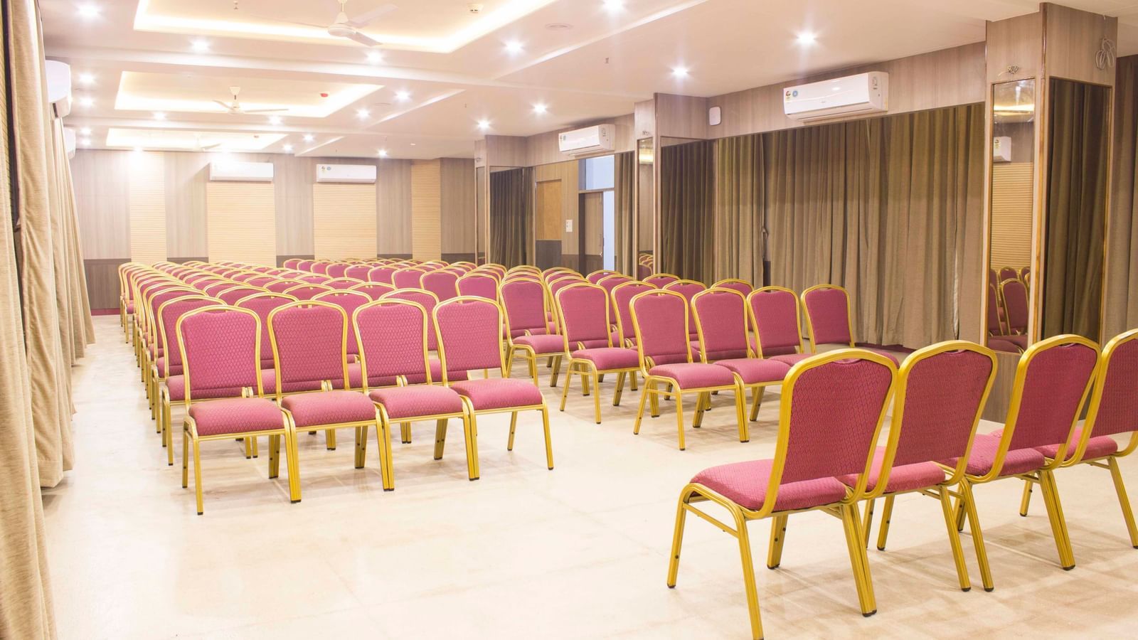 A wide shot of a banquet hall with multiple rows of empty pink and gold chairs. | Banquet Hall | Daksh The Madhuvan Suites