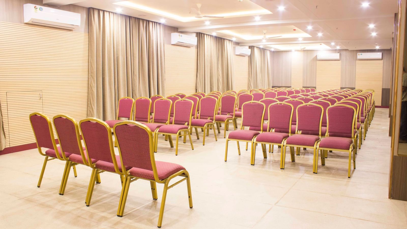 A wide shot of a banquet hall with multiple rows of empty pink and gold chairs. | Banquet Hall | Daksh The Madhuvan Suites