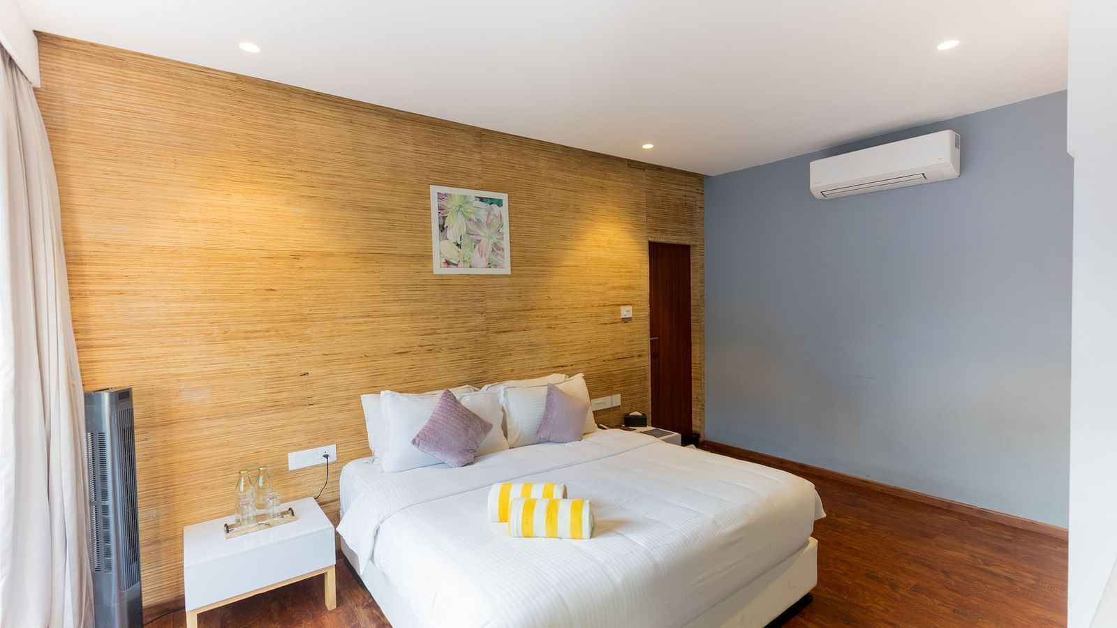 Grand Pool Villa at Darza Resorts features a white bed, a distinctive wooden-panelled accent wall, and a light blue wall with an air conditioning unit. 
