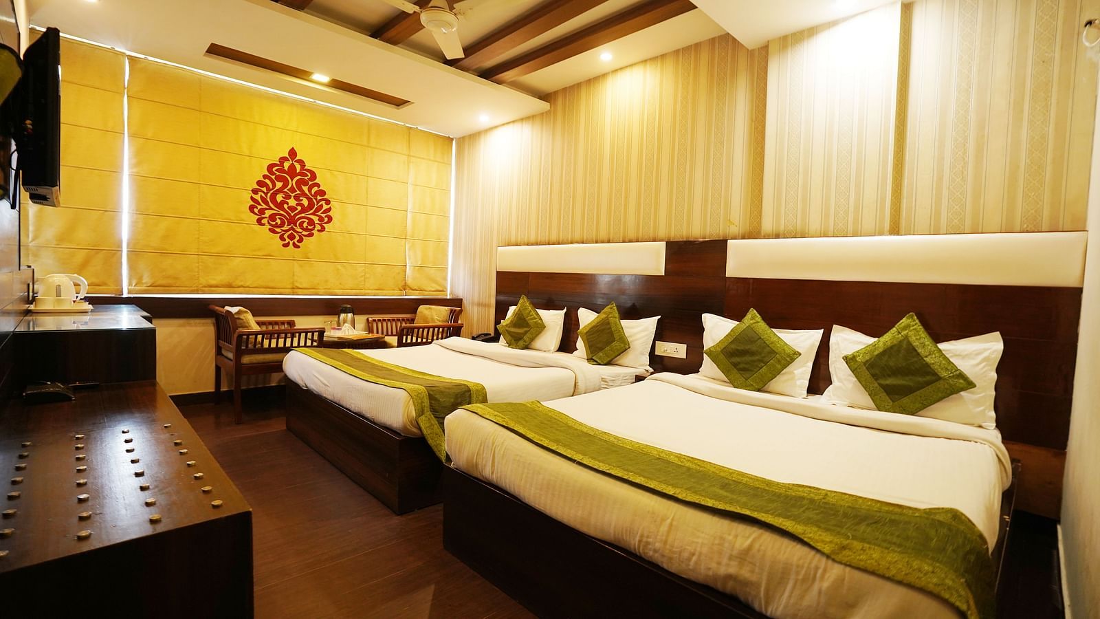 A spacious hotel room with two beds, one queen and one twin, dressed in white linens with green accents, a dark wood dresser, a yellow decorative wall hanging, and a small seating area - Diamond Hotels