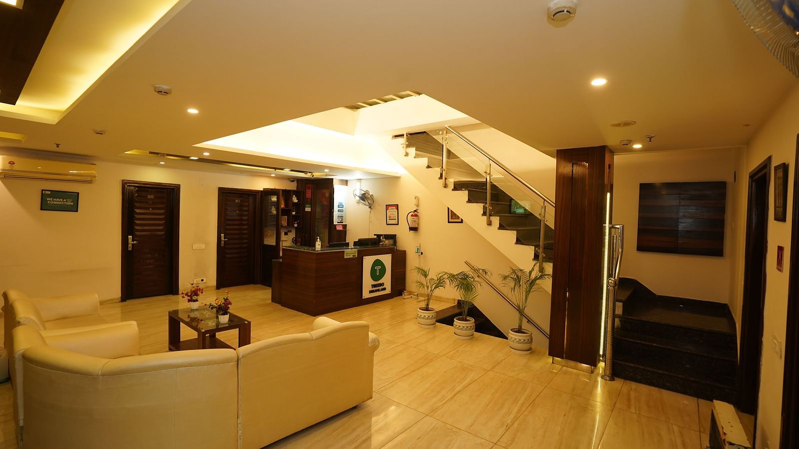 A hotel lobby with a curved white sofa, a small coffee table, a staircase leading to an upper level, and a reception desk in the background - Diamond Hotels