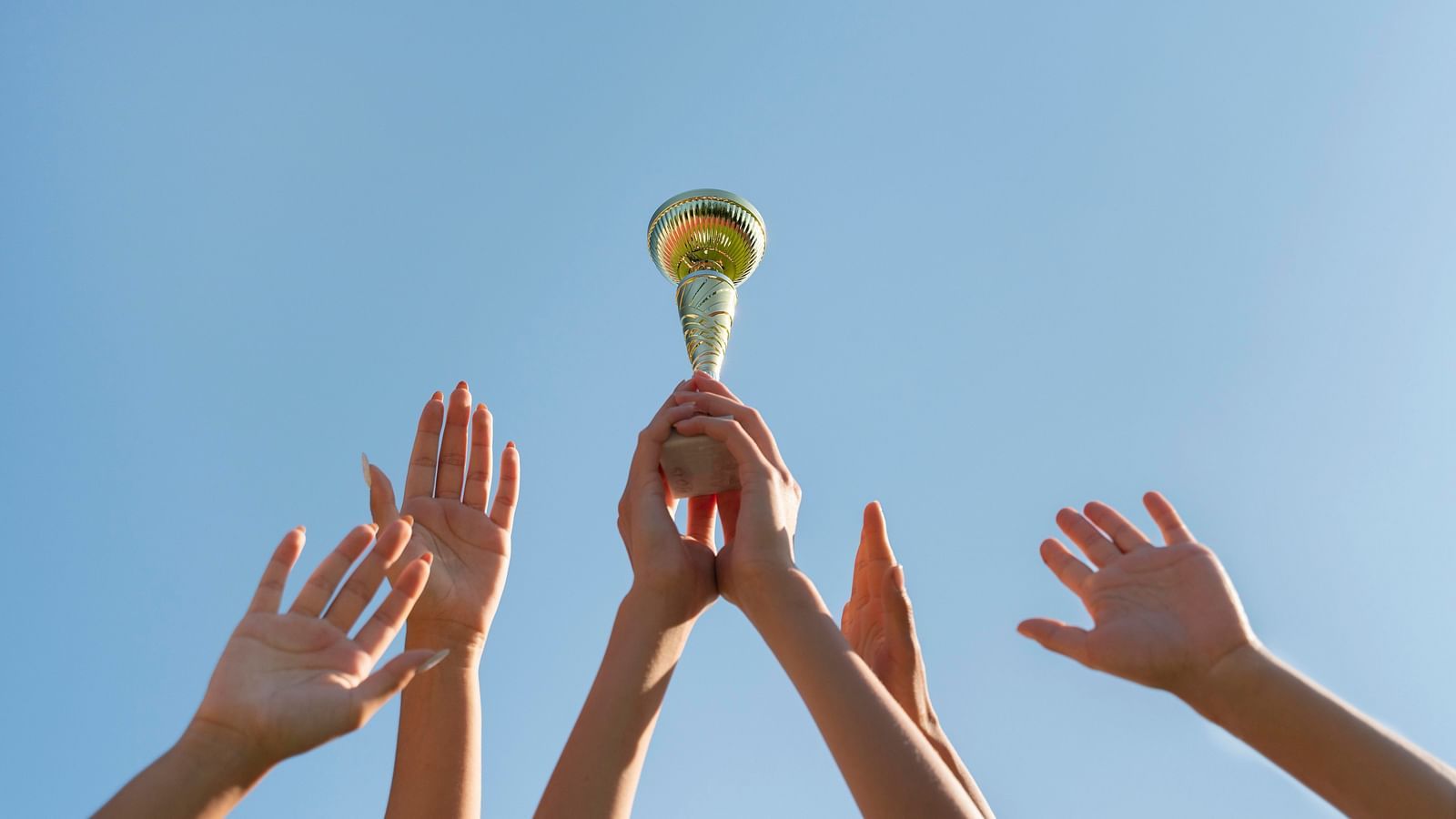 Multiple hands reaching towards a trophy held up against a clear blue sky, symbolising teamwork and achievement.