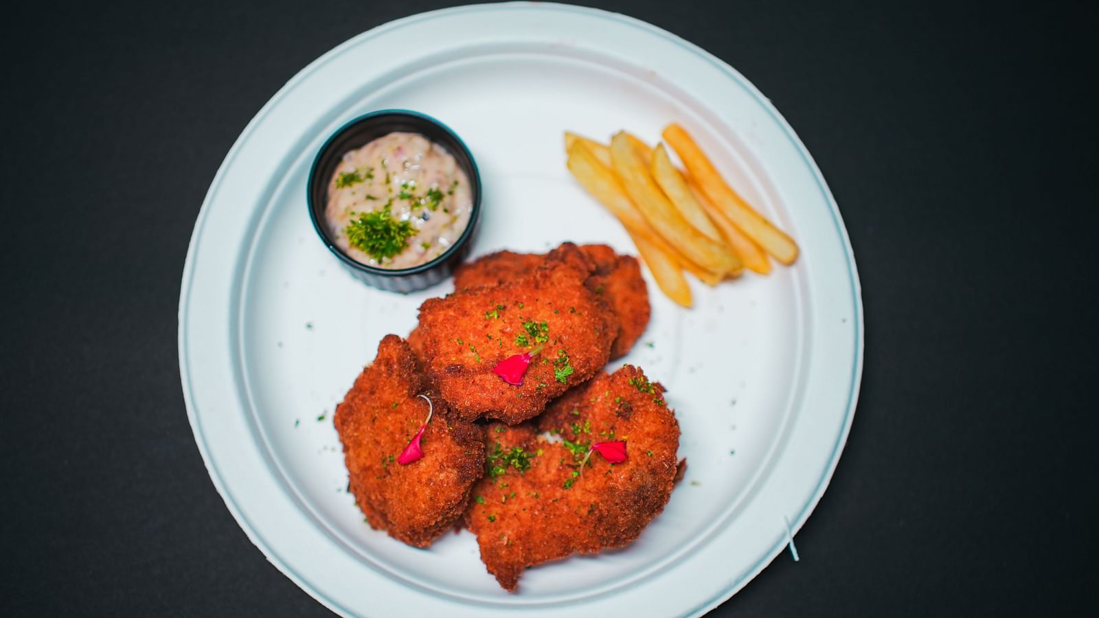 A white plate served with three pieces of crispy fried chicken, a side of golden French fries, and a small bowl of creamy dip, photographed on a dark background at D Café in Dip Holidays.