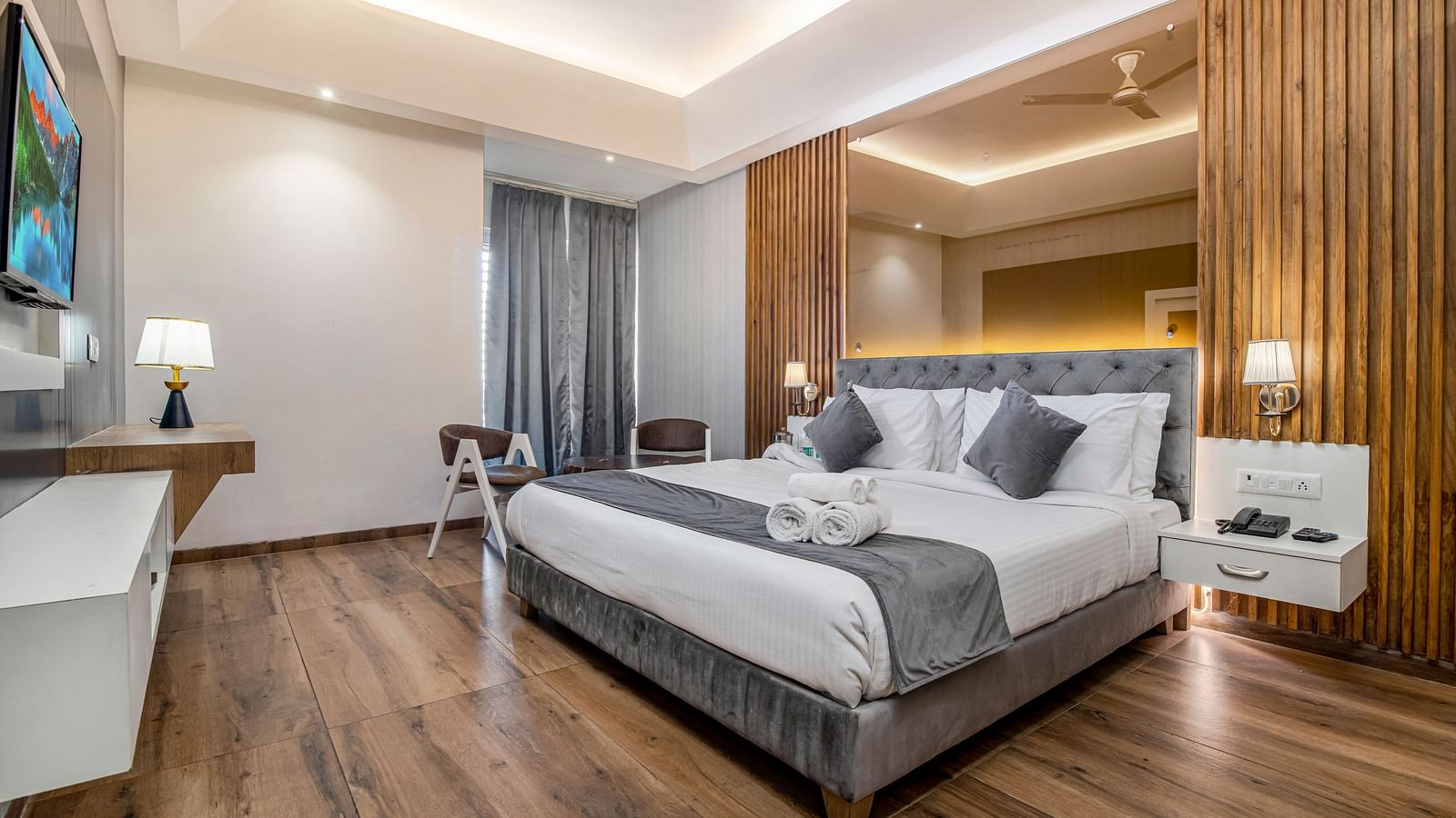 An image of the deluxe room featuring a bed, bed side tables, wall mounted flat screen TV, chairs and table, lamps and other room amenities - DLS Ganga Bliss, Haridwar