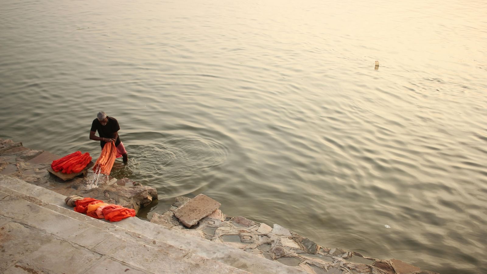 A person washing clothes on stone steps by a riverbank, with cloths placed nearby on the ground.
