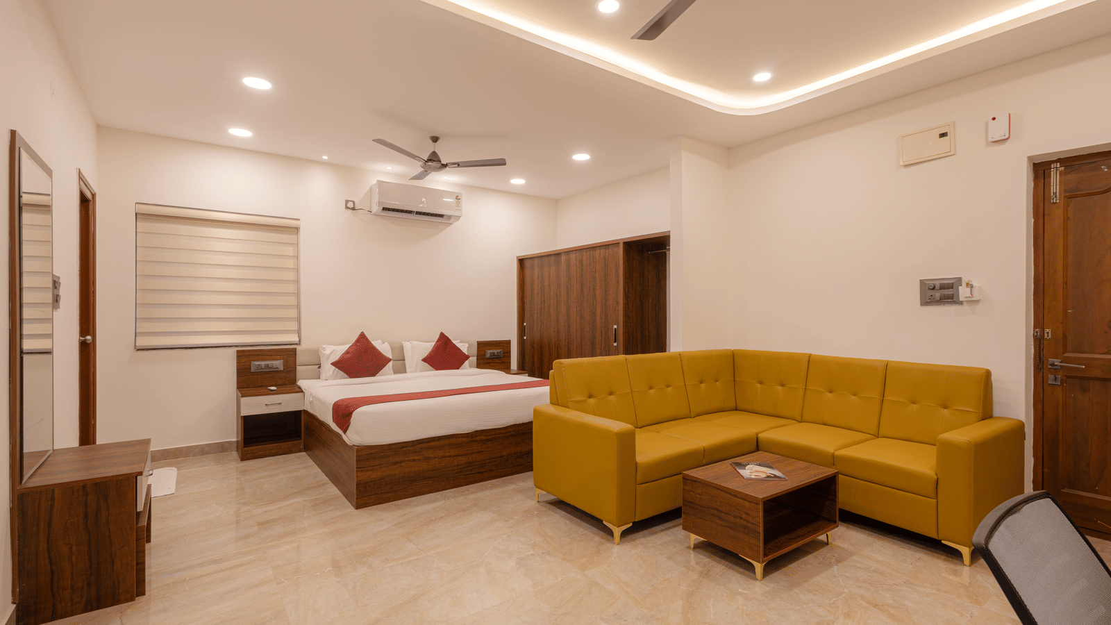 A room at Elysium Studio Suites, Madhapur, shows a bed, a yellow sofa, and a table on a tiled floor.
