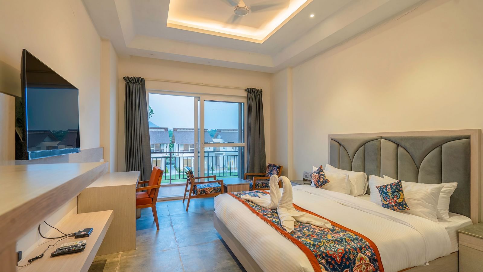 A spacious hotel room with a king size bed, a flat-screen TV, a work desk, and a balcony offering a scenic view of the surrounding landscape.