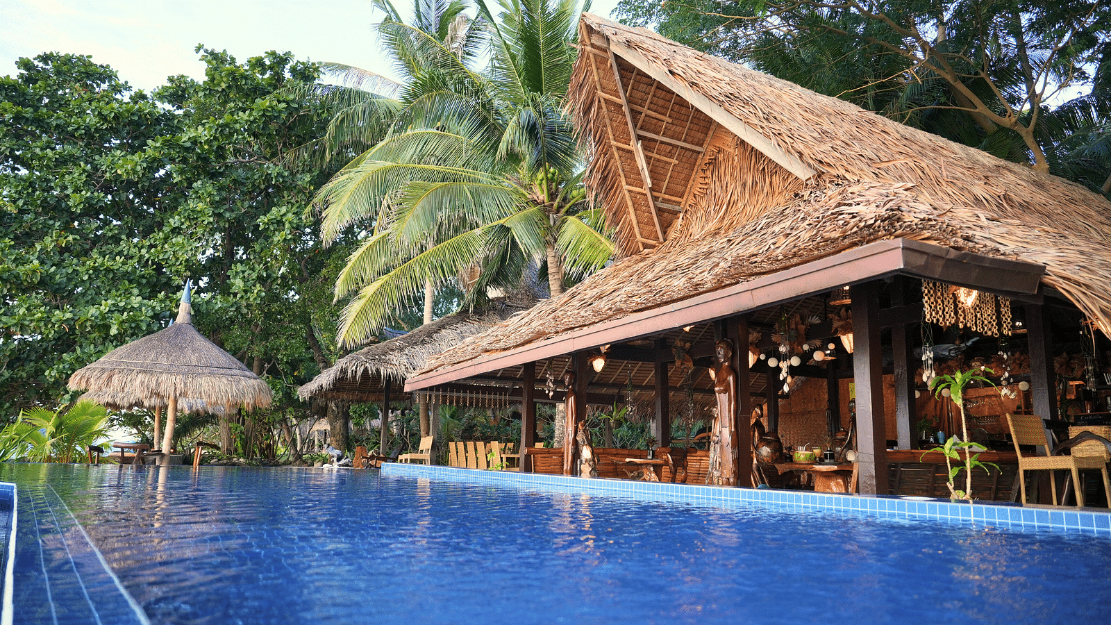 Poolside view with thatched-roof restaurant building and palm trees at Flower Island Resort.