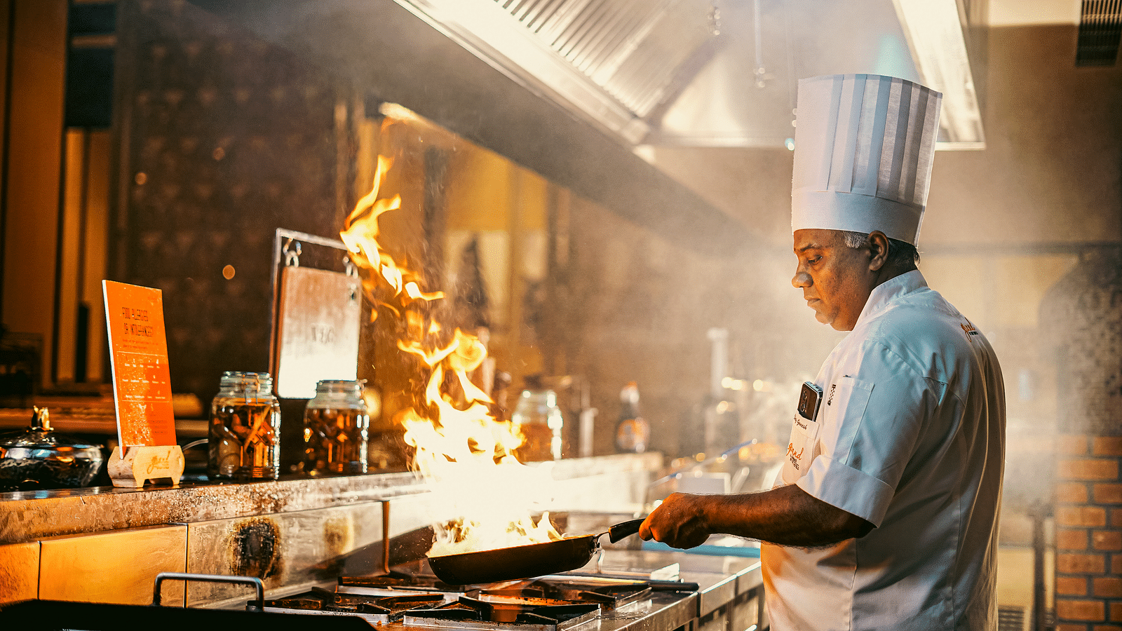 A chef expertly working at a live station, with flames rising from the pan, showcasing a vibrant culinary action - Grand Madurai by GRT Hotels
