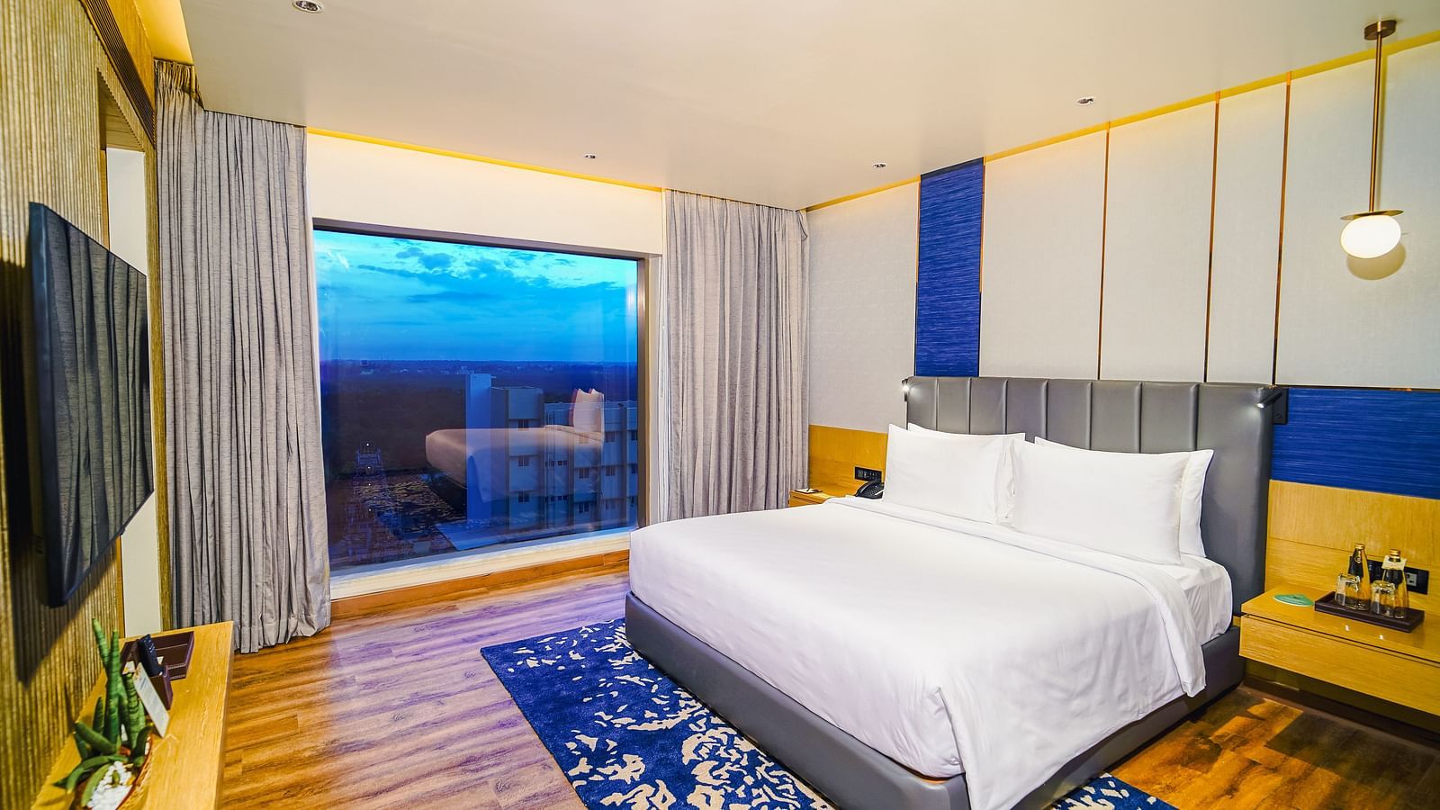 A Junior Suite in Madurai with a beautiful sunset view from the window, a blue carpet neatly arranged under the bed and a tv mounted on the wall - Grand Madurai by GRT Hotels