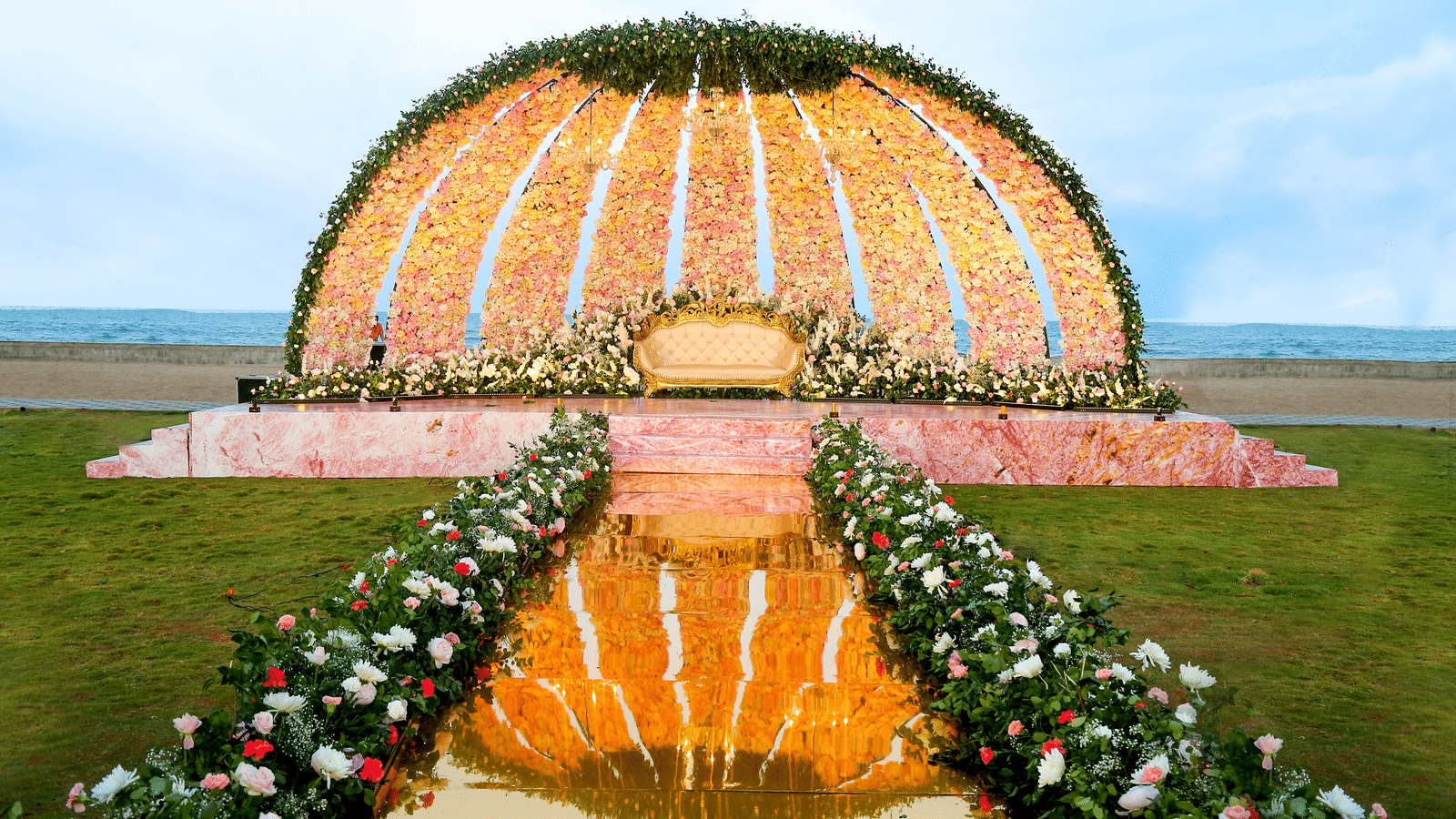 A large, decorative outdoor archway made of white and orange flowers and fabric, leading to a path on a beach at sunset - Grande Bay Resort & Spa, Mamallapuram