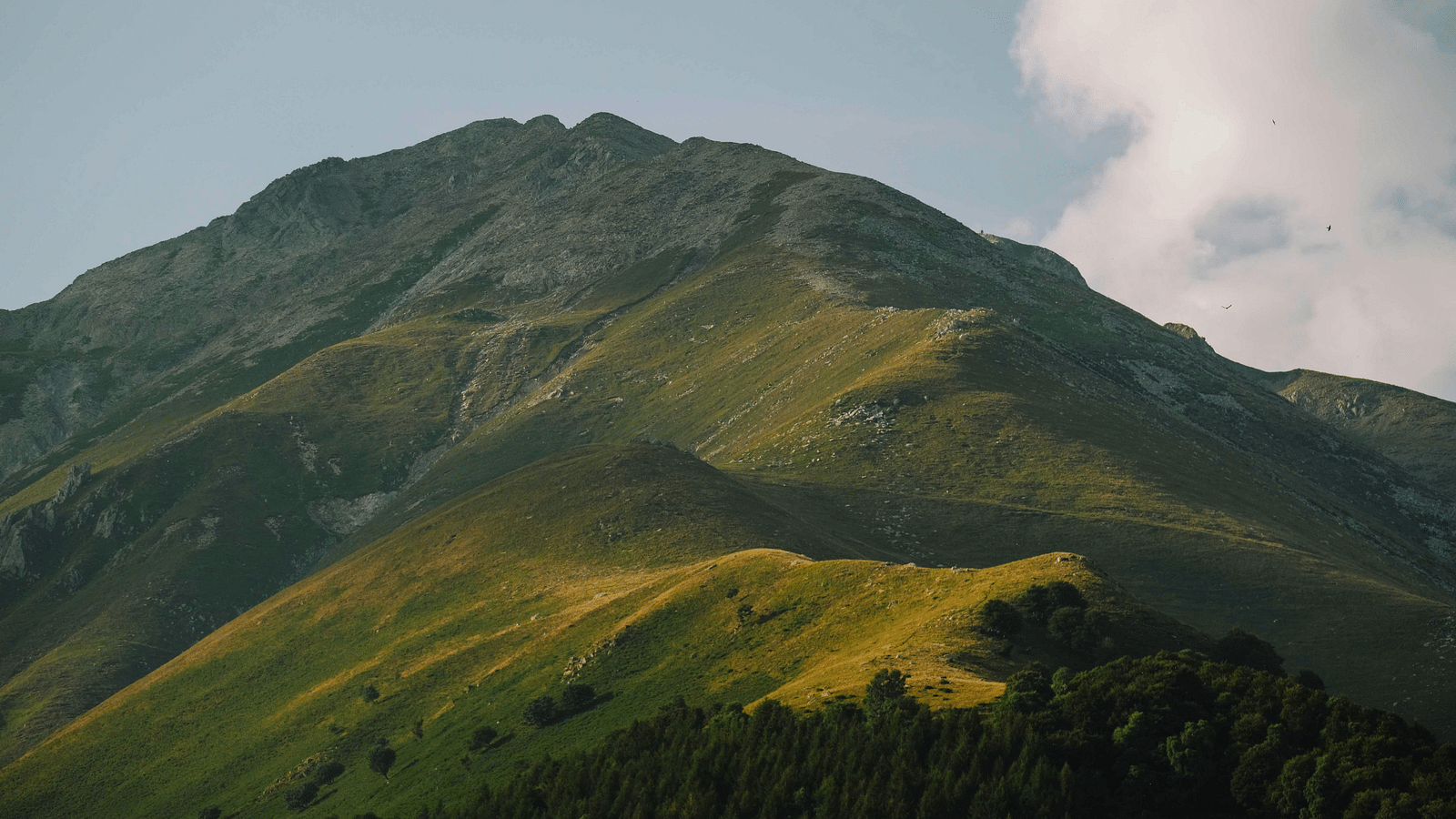 A picturesque view of a green mountain peak, with rolling hills covered in grass and a light cloud hovering in the blue sky.